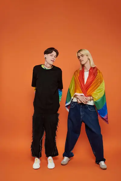 Deux jeunes amis, debout avec un drapeau arc-en-ciel, posent sur un fond orange. — Photo de stock