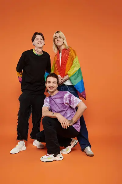Tres jóvenes amigos en trajes elegantes posan sobre un telón de fondo naranja. Uno de ellos sostiene una bandera del orgullo del arco iris. — Stock Photo