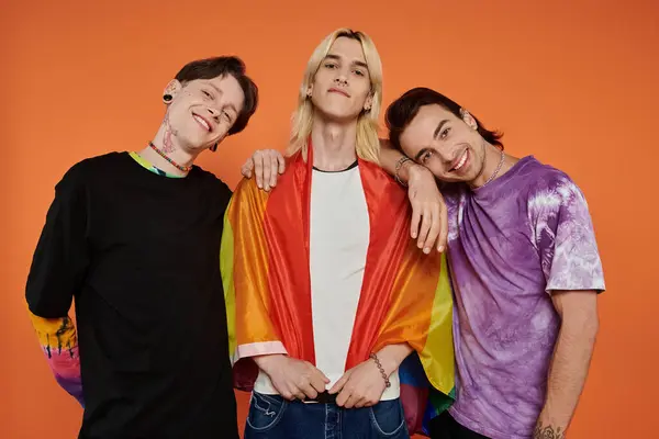 Tres jóvenes amigos con un atuendo elegante posan junto con una bandera de orgullo envuelta alrededor de uno de ellos, contra un fondo naranja. — Stock Photo