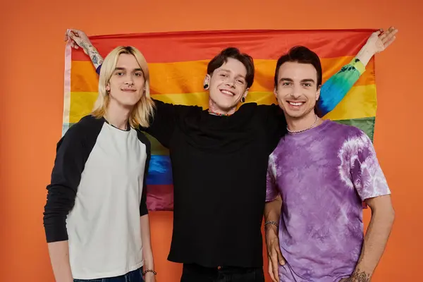 Tres jóvenes amigos se paran juntos frente a un fondo naranja, sosteniendo una bandera de arco iris y sonriendo. — Stock Photo