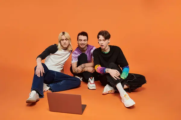 Three young queer friends in vibrant attire are seated on an orange backdrop, watching something on a laptop. — Stock Photo