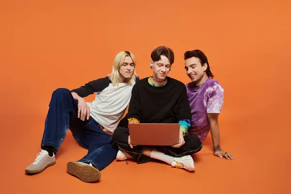 Three young queer friends in stylish, colorful clothing sit on a bright orange background, one using a laptop. — Stock Photo