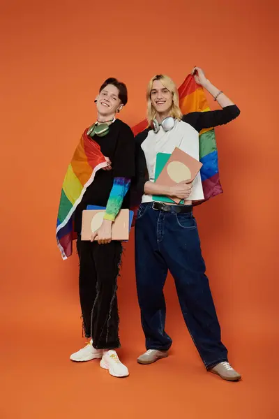 Dos jóvenes amigos queer se levantan orgullosamente con banderas de arco iris envueltas alrededor de sus hombros, sosteniendo libros sobre un fondo naranja brillante. — Stock Photo