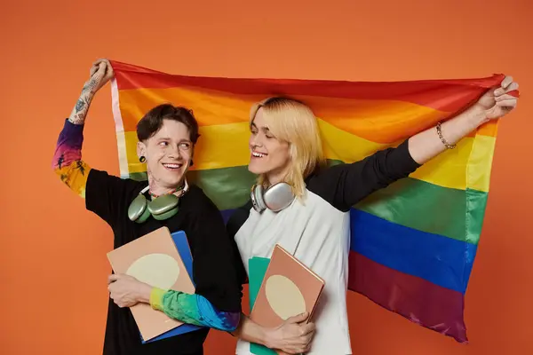 Dos jóvenes amigos sostienen una bandera de orgullo y posan con cuadernos frente a un fondo naranja. — Stock Photo