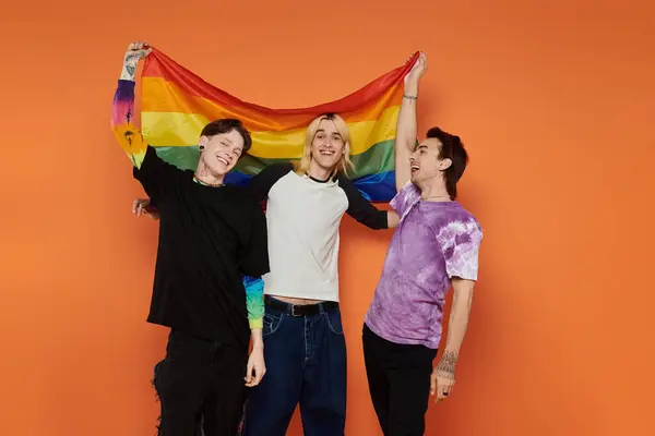 Tres jóvenes amigos, vestidos con un atuendo elegante y vibrante, posan sobre un fondo naranja mientras sostienen una bandera de arco iris. — Stock Photo
