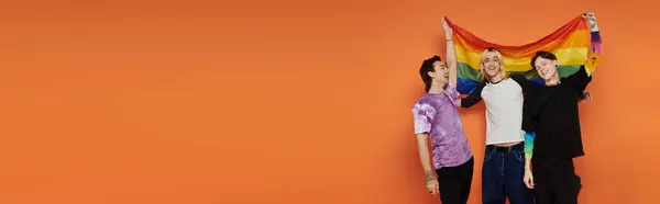 Tres jóvenes amigos sostienen una bandera del orgullo del arco iris frente a un fondo naranja. — Stock Photo