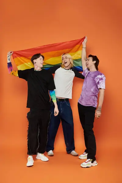 Trois jeunes amis en tenue élégante posent ensemble tenant un drapeau de fierté arc-en-ciel devant un fond orange. — Photo de stock