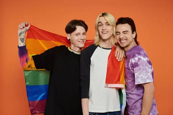 Trois jeunes amis se tiennent ensemble, souriant et tenant un drapeau de fierté arc-en-ciel, célébrant la diversité et l'inclusion. — Photo de stock