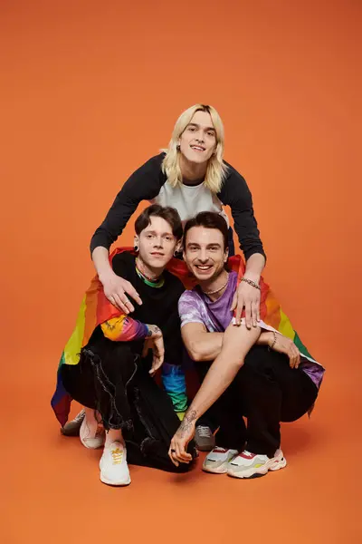 Tres amigos se paran frente a un vibrante telón de fondo naranja, envueltos en una bandera de orgullo, mostrando su unidad y solidaridad LGBTQIA. — Stock Photo