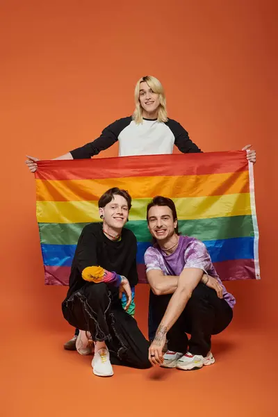 Tres jóvenes amigos, dos arrodillados y uno de pie, sosteniendo una bandera del orgullo del arco iris sobre un fondo naranja. — Stock Photo