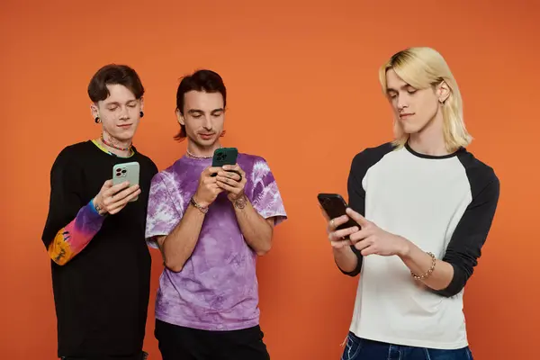 Three LGBTQIA friends stand against orange backdrop, each on their phones, dressed in stylish clothing. — Stock Photo
