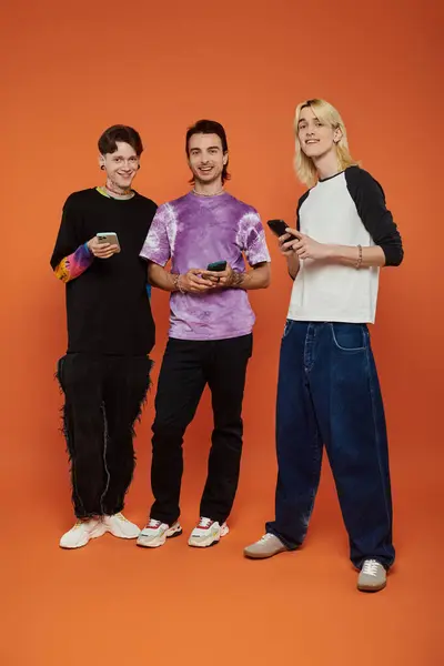 Three young queer friends stand in front of an orange background, each wearing colorful and stylish outfits. — Stock Photo