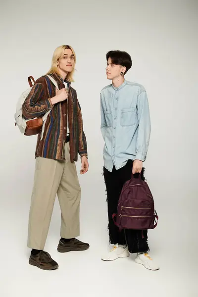 Dos jóvenes estudiantes queer con atuendo casual se paran con mochilas frente a un fondo gris. — Stock Photo