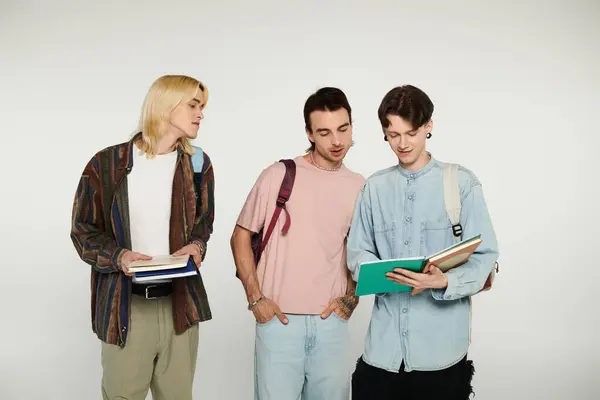Three young men in casual clothing stand together on a grey background, one reading a book while the others look on. — Stock Photo