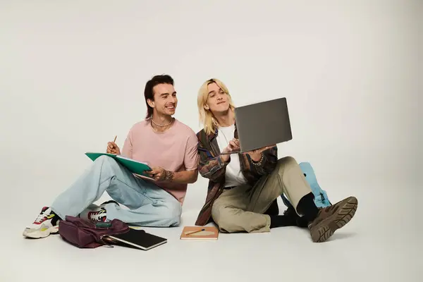 Two young queer students work on a project together, one using a laptop while the other takes notes. — Stock Photo