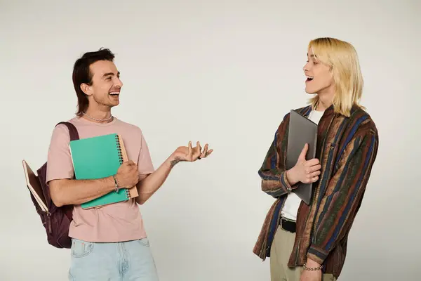 Dois jovens estudantes gays estão juntos, um segurando um caderno e outro um laptop, compartilhando uma risada contra um fundo cinza. — Fotografia de Stock