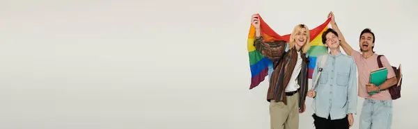 Tres jóvenes estudiantes se paran juntos, sonriendo y sosteniendo una bandera de orgullo del arco iris. — Stock Photo