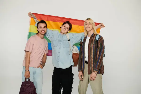 Tres jóvenes estudiantes queer se paran juntos, sosteniendo una bandera de arco iris frente a un fondo gris. — Stock Photo