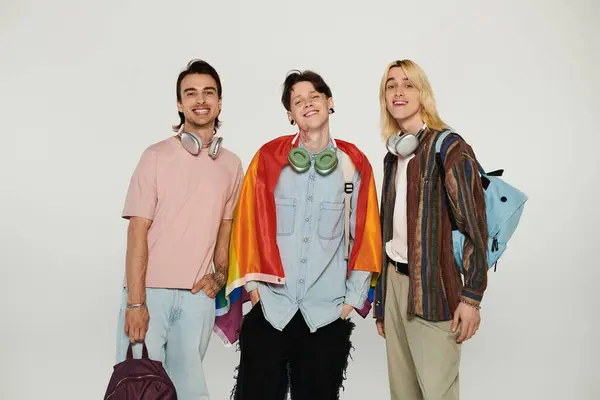 Tres jóvenes estudiantes, parte de la comunidad LGBTQIA, se paran frente a un fondo gris, sonriendo y sosteniendo una bandera de orgullo. — Stock Photo