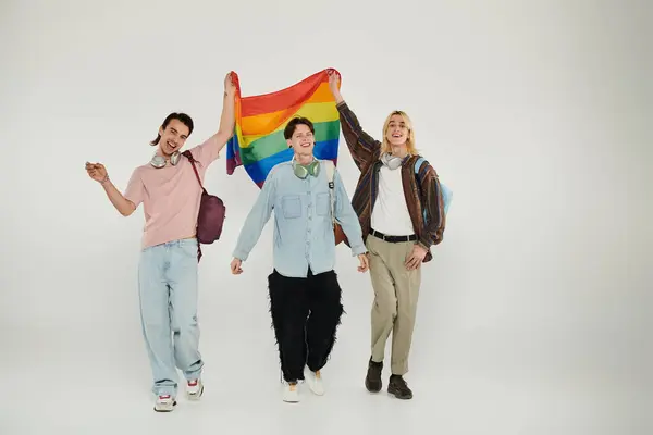 Tres jóvenes estudiantes con ropa casual sostienen una bandera de arco iris frente a un fondo blanco. — Stock Photo