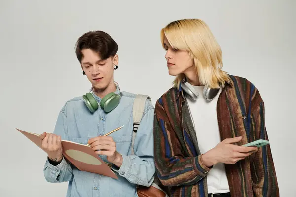 Two young queer students stand together in casual attire, one taking notes and the other looking on. — Stock Photo