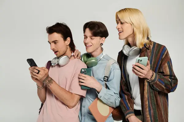 Three young queer students stand together, looking at phones and laughing. — Stock Photo