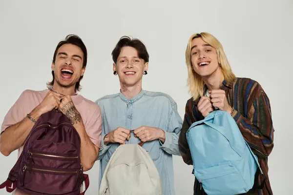 Three young, queer students in casual attire pose with their backpacks against a gray background. — Stock Photo