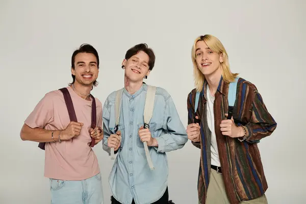 Três jovens estudantes queer ficar juntos, sorrindo, vestindo roupas casuais e mochilas contra um fundo cinza. — Fotografia de Stock