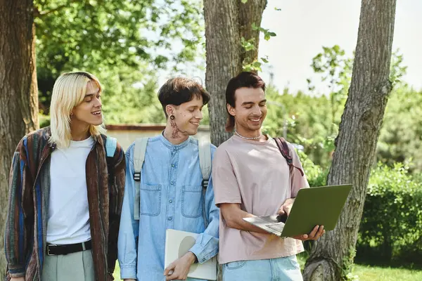 Tre giovani amici queer, vestiti con disinvoltura, stanno insieme in un parco, ridendo e condividendo un computer portatile. — Foto stock