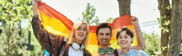 Three young queer friends stand together outdoors, smiling and holding a rainbow pride flag. — Stock Photo