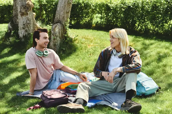 Two young queer friends sit on a blanket in a grassy park, laughing and enjoying each others company. — Stock Photo
