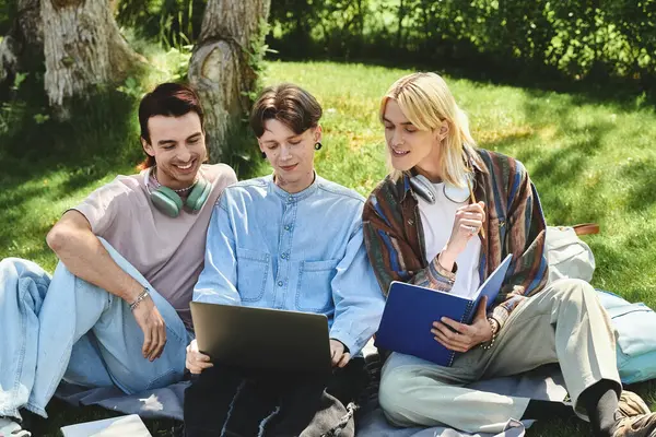Three young queer friends sit together in a grassy park, looking at a laptop and studying. — Stock Photo