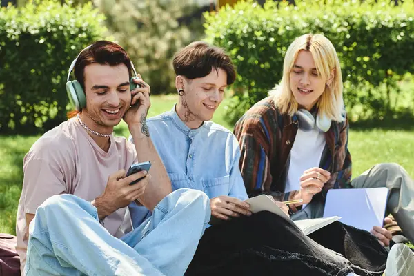 Three young queer friends enjoy a chill day on the grass: one with headphones, one reading, and the last on their phone. — Stock Photo