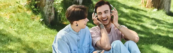 Two young friends, casually dressed, sit on a grassy lawn and listen to music on headphones. — Stock Photo