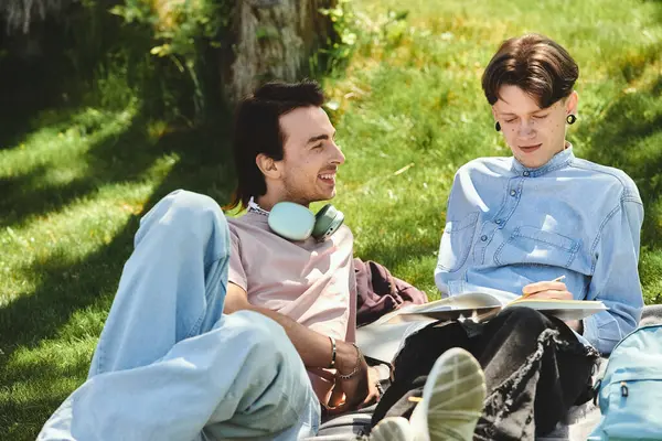 Two young queer friends relax on a blanket in a park, one studying while the other looks on with a smile. — Stock Photo