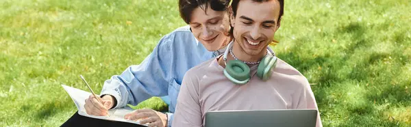 Zwei junge queere Freunde sitzen im Gras, einer arbeitet am Laptop, der andere schreibt auf Papier. — Stockfoto