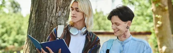 Two young friends, dressed in casual attire, stand outdoors under a tree, with one reading a book. — Stock Photo