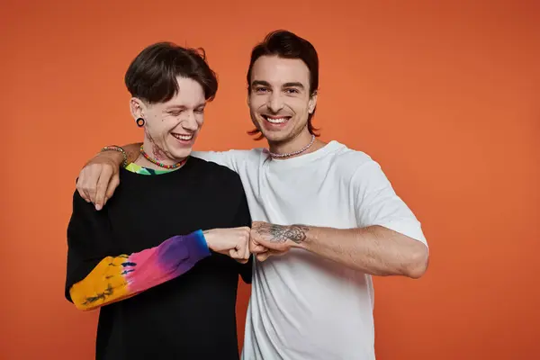 Two young men wearing colorful attire bump fists while laughing on an orange background. — Stock Photo