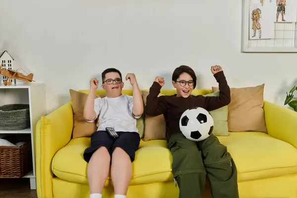 Two boys, one with Down syndrome, cheer on their favorite team while sitting on a yellow couch. — Stock Photo