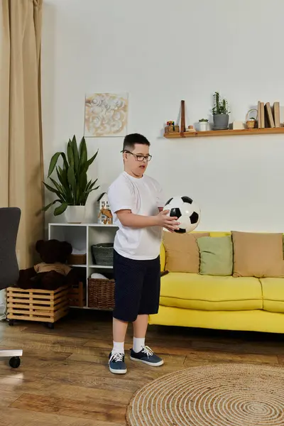 Um menino com síndrome de Down segura uma bola de futebol em sua sala de estar. — Fotografia de Stock