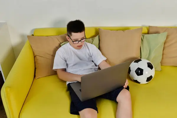 A young boy with Down syndrome sits on a yellow couch at home, focused on his laptop. — Stock Photo