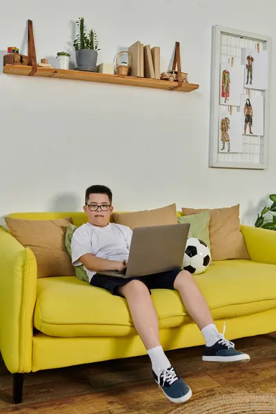 A young boy with Down syndrome sits on a yellow couch, using a laptop. — Stock Photo