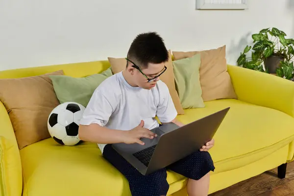 A young boy with Down syndrome sits on a yellow couch using a laptop. — Stock Photo