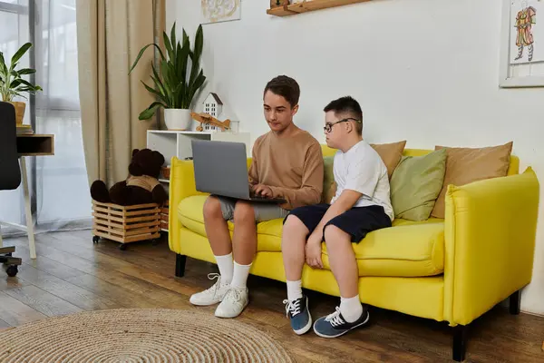 Dois meninos, um com síndrome de Down, estão sentados em um sofá amarelo em casa, usando um laptop. — Stock Photo