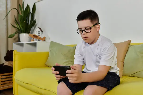 A young boy with Down syndrome sits on a yellow couch, his eyes glued to his phone. — Stock Photo