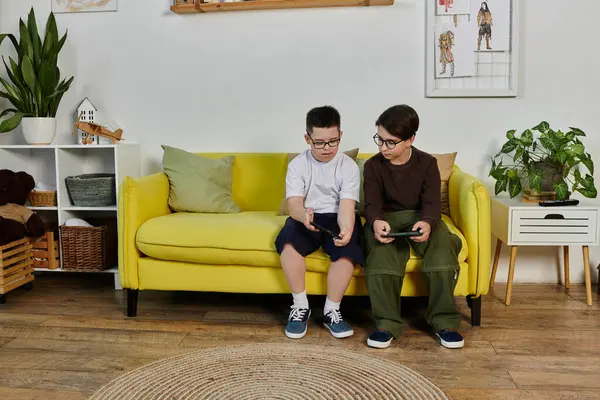 Two young boys, one with Down syndrome, sit on a yellow sofa and play video games. — Stock Photo