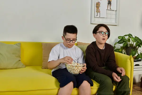 Two boys, one with Down syndrome, sit on a yellow couch together, enjoying popcorn. — Stock Photo