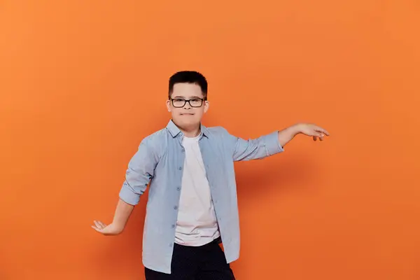 Boy with Down syndrome in glasses poses smiling with arms outstretched against orange. — Stock Photo