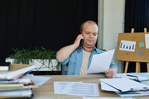 Un hombre con inclusividad se sienta en un escritorio, revisa documentos y habla por teléfono. - foto de stock