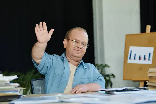 Человек с инклюзивностью в очках сидит за столом, улыбаясь и поднимая руку в приветствии. — стоковое фото